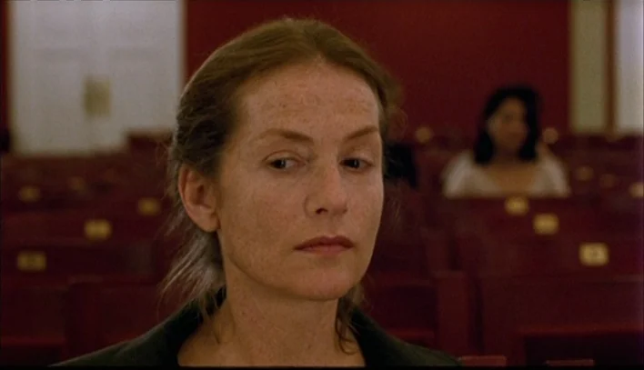 The Piano Teacher (2001) movie still: closeup — Rows of empty red auditorium seats fill the dimly lit space. A woman is in the foreground with…