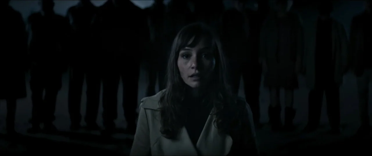 Offseason (2022) movie still: medium — A woman stands in an open area at night surrounded by a large crowd. A group of dark, silhouetted figures…