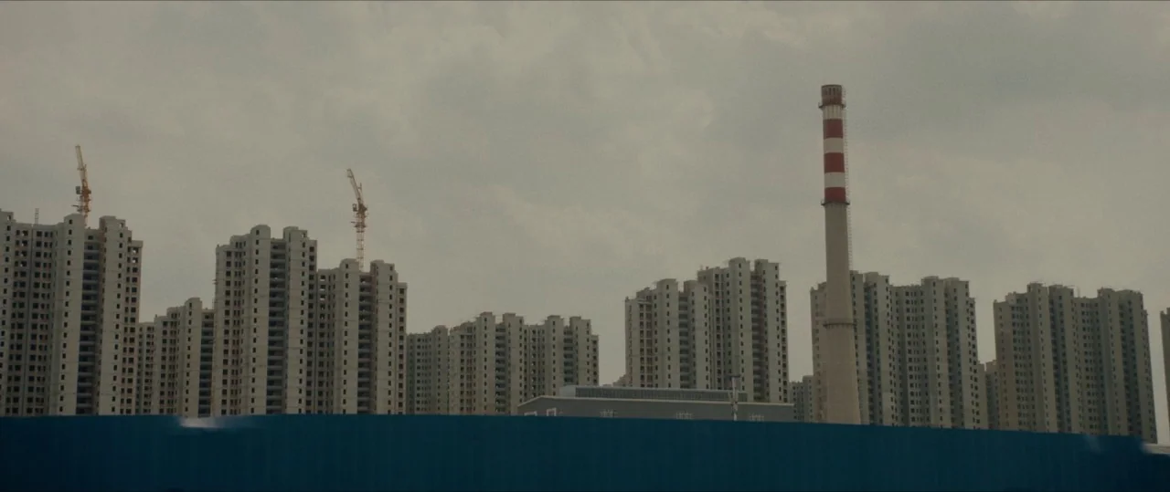 The Farewell (2019) movie still: establishing — A dense cluster of unfinished high-rise concrete apartment blocks fills the background under grey…