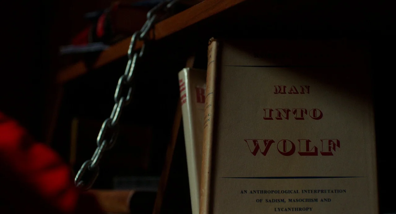 My Animal (2023) movie still: closeup — A hardcover book with the title Man Into Wolf sits on a dimly lit shelf. A metal chain hangs suspended in…