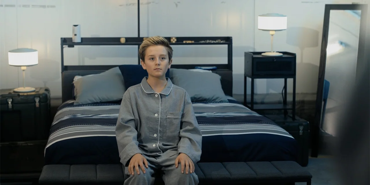 Playdate (2025) movie still: medium — A modern, minimalist bedroom features a dark striped bedspread and two identical bedside lamps. A young boy in…