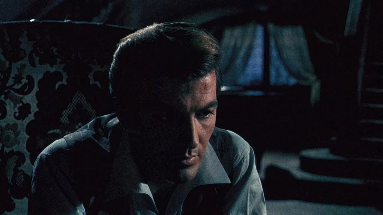 The Whip and The Body (1963) movie still: closeup — A darkened interior space features a man seated in an ornate, high-backed patterned armchair. A…