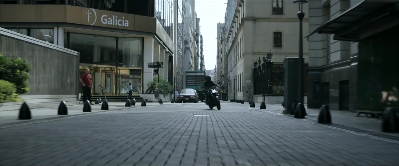 Checkmate (2015) movie still: wide — A wide city street features a cobblestone surface and a large bank office building. A person rides a motorcycle…