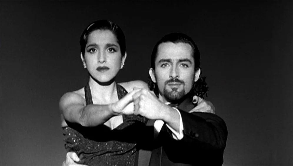 The Tango Lesson (1997) movie still: medium — A man and woman stand close together against a pitch black background. They hold their hands together…
