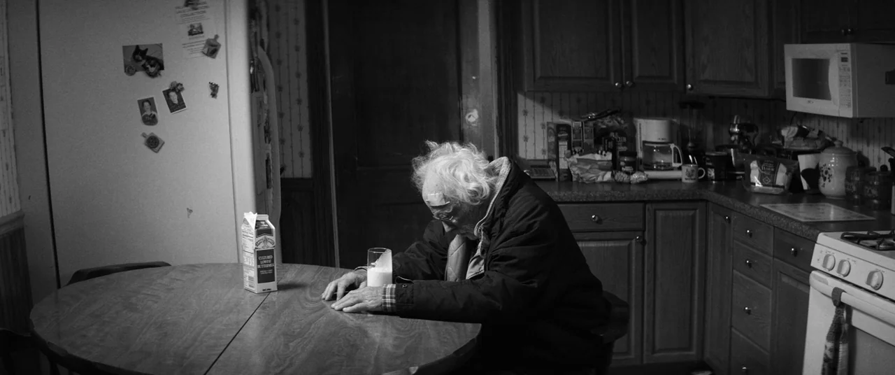 Nebraska (2013) movie still: medium — A dark, residential kitchen with wooden cabinets and wallpaper featuring small patterns. An elderly man with a…