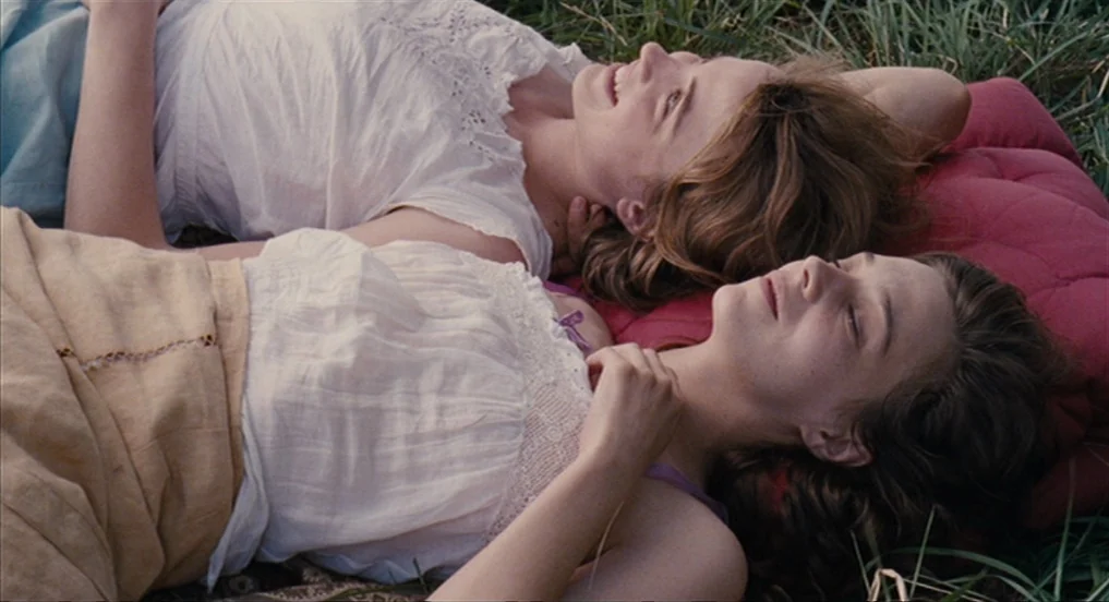 House of Tolerance (2011) movie still: medium — Two young women recline on a red cushion nestled within a grassy outdoor field. They lie close…