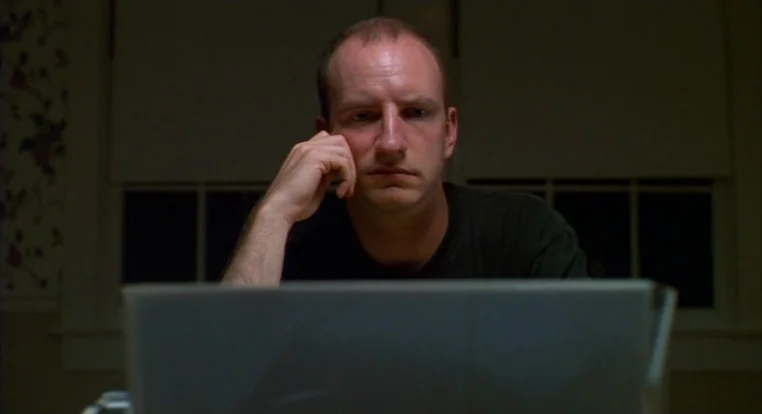 Schizopolis movie still: medium — A man sits at a desk in a darkened room at night. He stares intently at the glowing screen of a laptop.