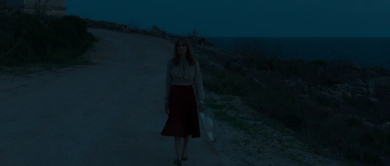By The Sea (2015) movie still: fullbody — A woman walks down a dirt road beside the sea during twilight hours. The landscape is sparse with low…