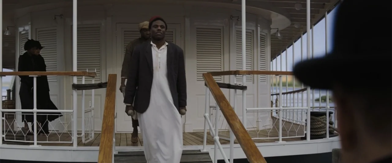Death on the Nile (2022) movie still: fullbody — The exterior deck of a white-painted wooden steamboat has railings and stairs. A man in a white…
