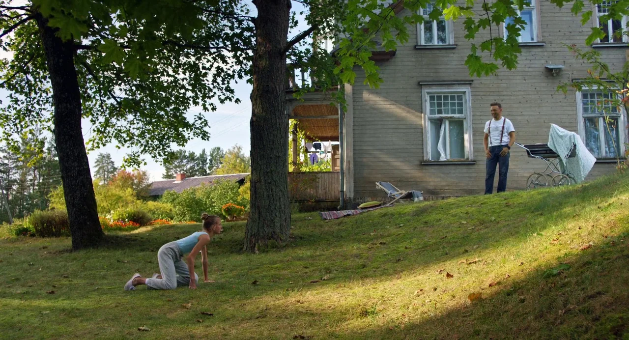 Hatching (2022) movie still: wide — A rural backyard features a wooden house with a porch and overgrown grass slopes. A woman crawls on all fours…