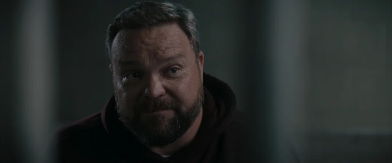 A Christmas Mystery (2022) movie still: closeup — A dark, dimly lit room with blurred vertical bars in the foreground. A man in a dark hoodie sits…