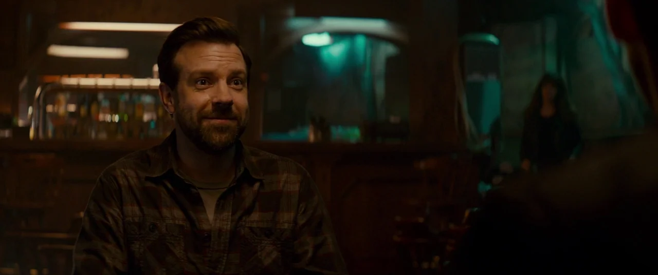 Colossal (2016) movie still: medium — A dark, dimly lit dive bar features a wooden counter and blurry background bottles. A man sits at a table…