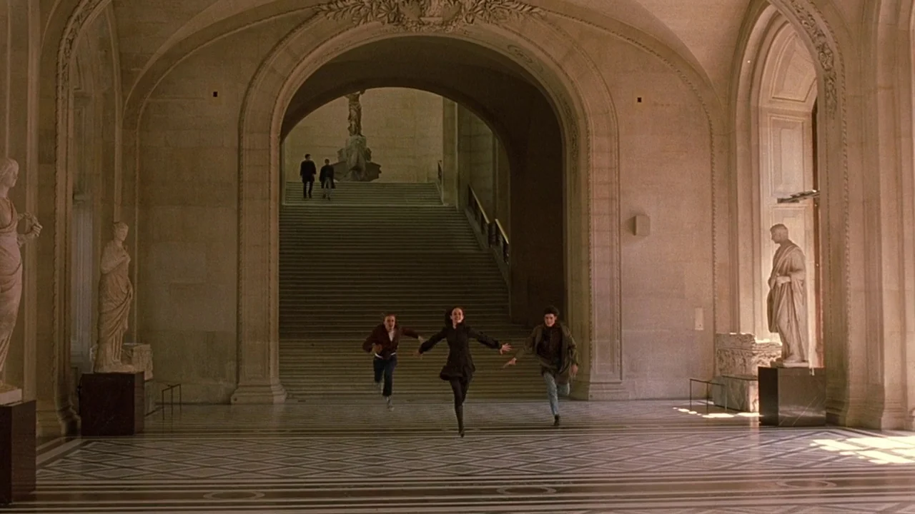 The Dreamers (2003) movie still: wide — A grand, high-ceilinged stone gallery features a wide staircase leading to an arched doorway. Three young…