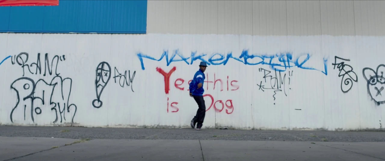 Da Sweet Blood of Jesus (2014) movie still: wide — A white corrugated metal wall covered in colorful spray-painted graffiti stands on asphalt…