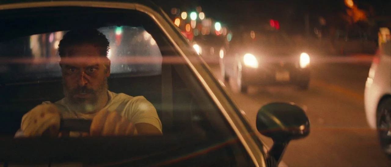 Archenemy (2020) movie still: medium — A man drives a car at night through a busy urban street with traffic. Blurred orange street lights and…