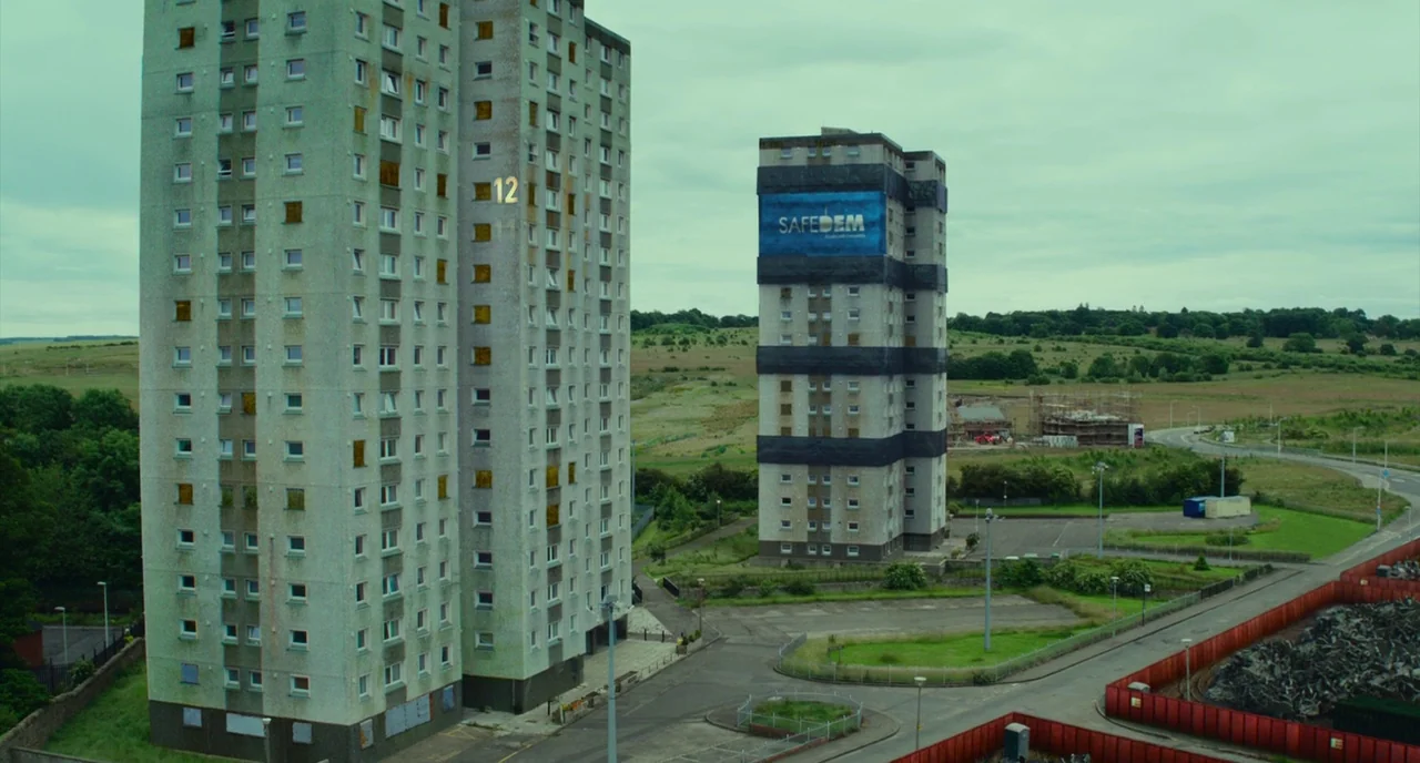 T2 Trainspotting (2017) movie still: establishing — Two tall apartment towers stand in a sparse, green field under a cloudy sky. One tower features…
