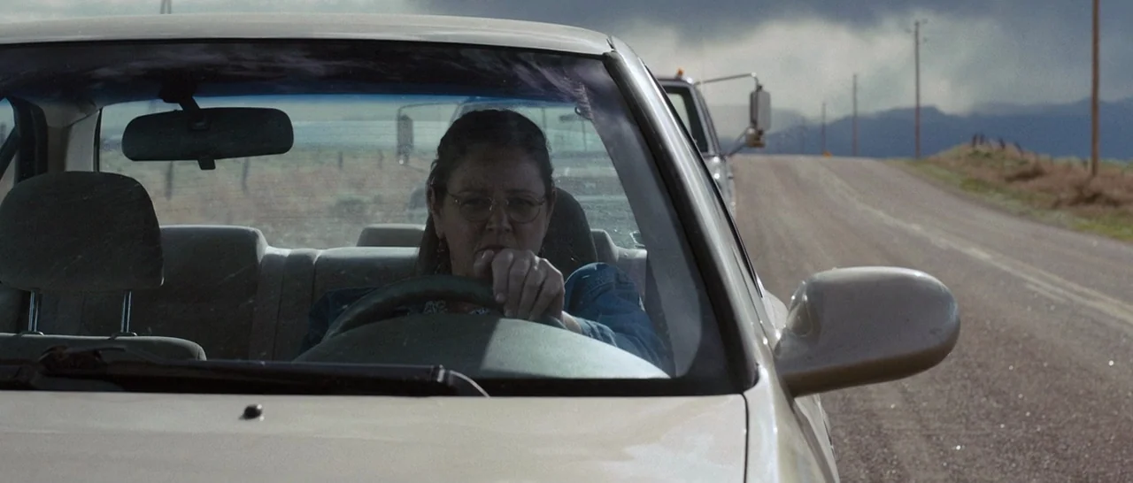 Cop Car (2015) movie still: medium — A woman drives a light-colored sedan on a paved road through open, dry landscape. A large pickup truck follows…