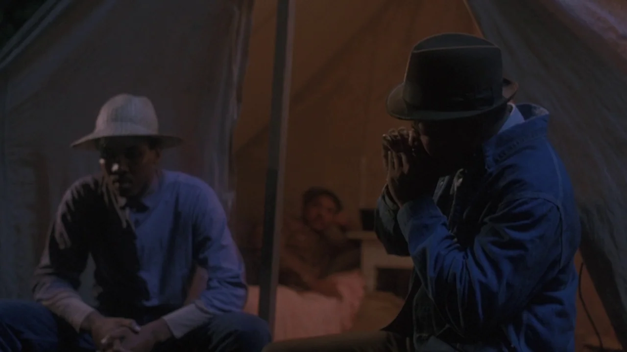 Matewan (1987) movie still: medium — A dimly lit canvas tent contains two men sitting together in the dark. One man plays a small musical instrument…