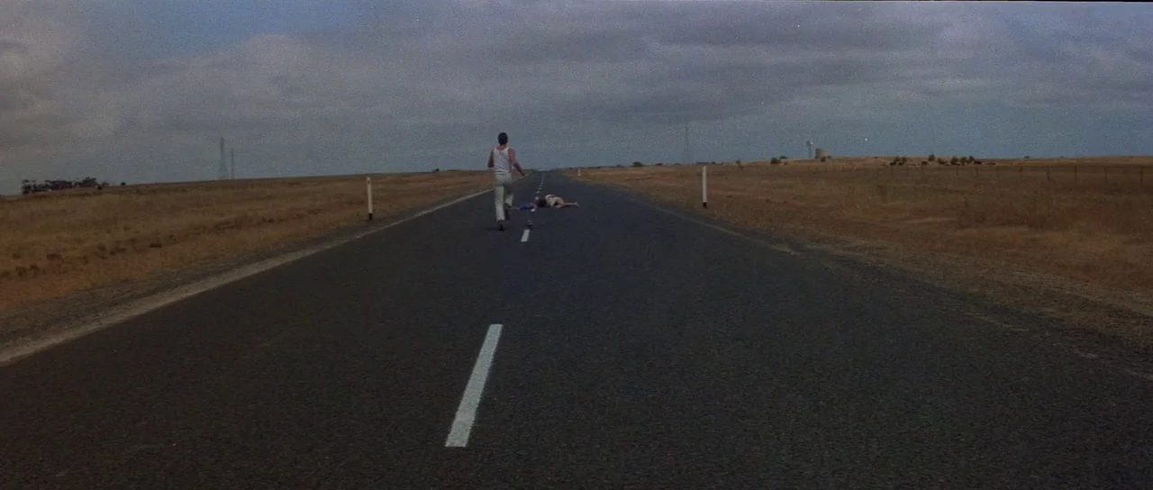 Mad Max (1979) movie still: wide — An asphalt road stretches through a vast, dry, and flat rural landscape under clouds. A lone man runs forward…