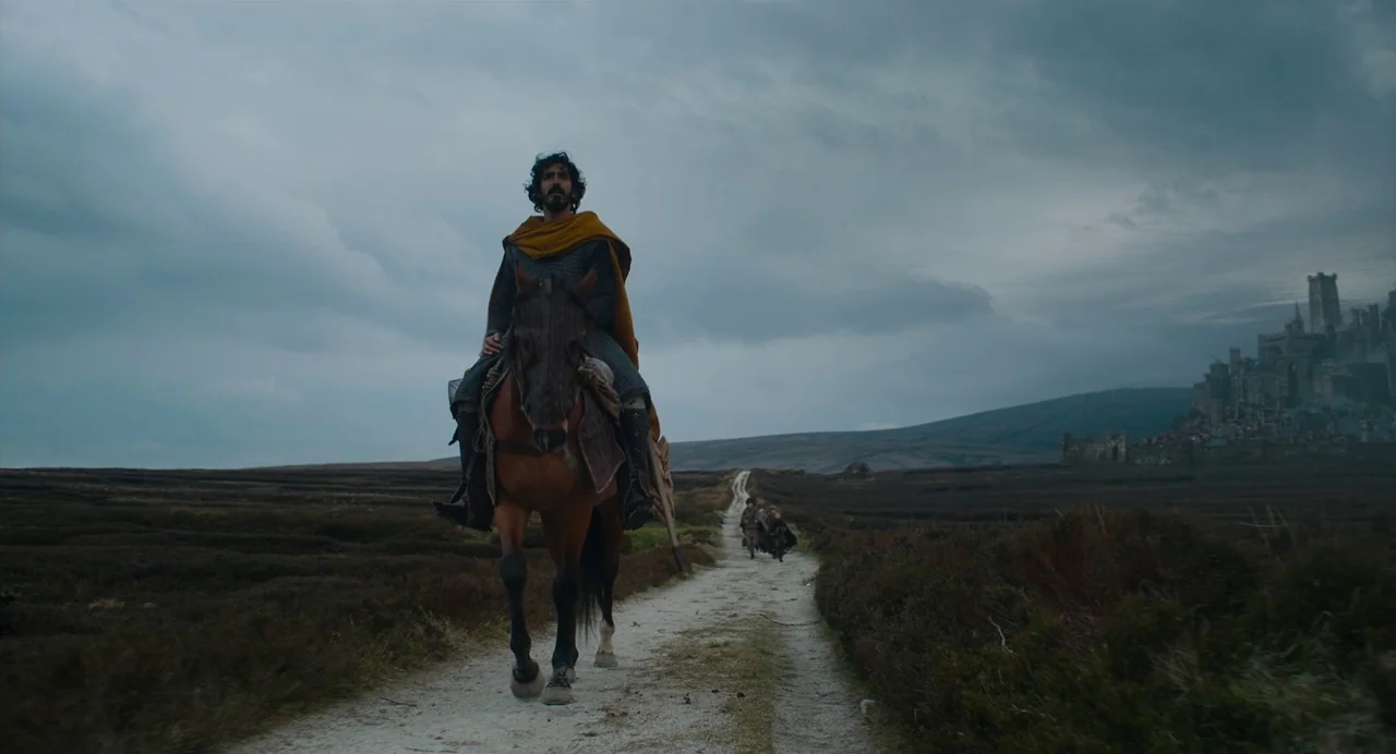The Green Knight (2021) movie still: fullbody — An open moorland features a dirt road leading toward a large stone citadel. A lone rider on…
