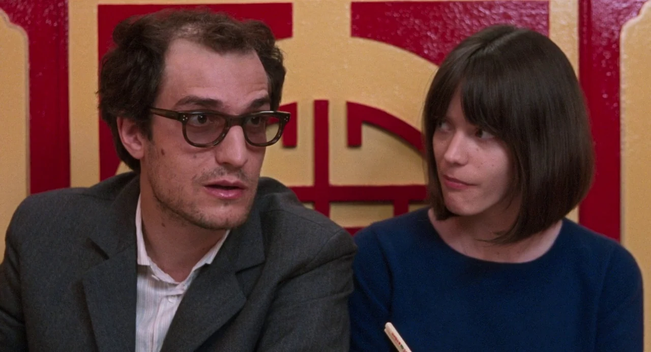 Godard Mon Amour (2015) movie still: medium — Two individuals sit in front of a red and gold decorative patterned wall panel. The man in glasses…