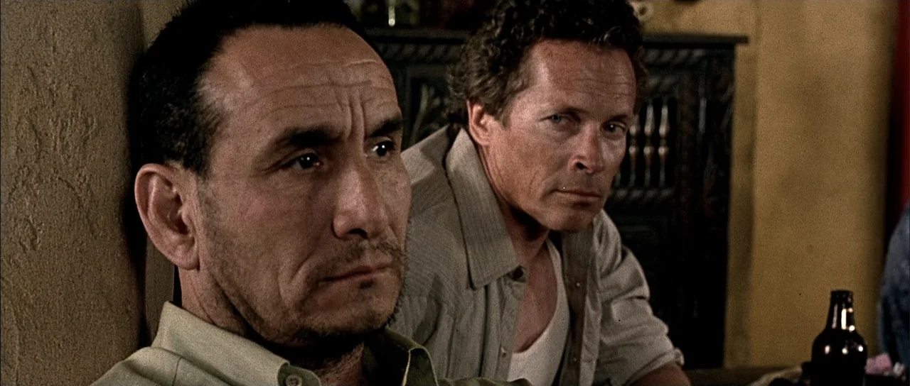 A Man Apart (2003) movie still: medium — A dimly lit interior space with textured walls and a wooden cabinet visible behind. Two men are seated at a…