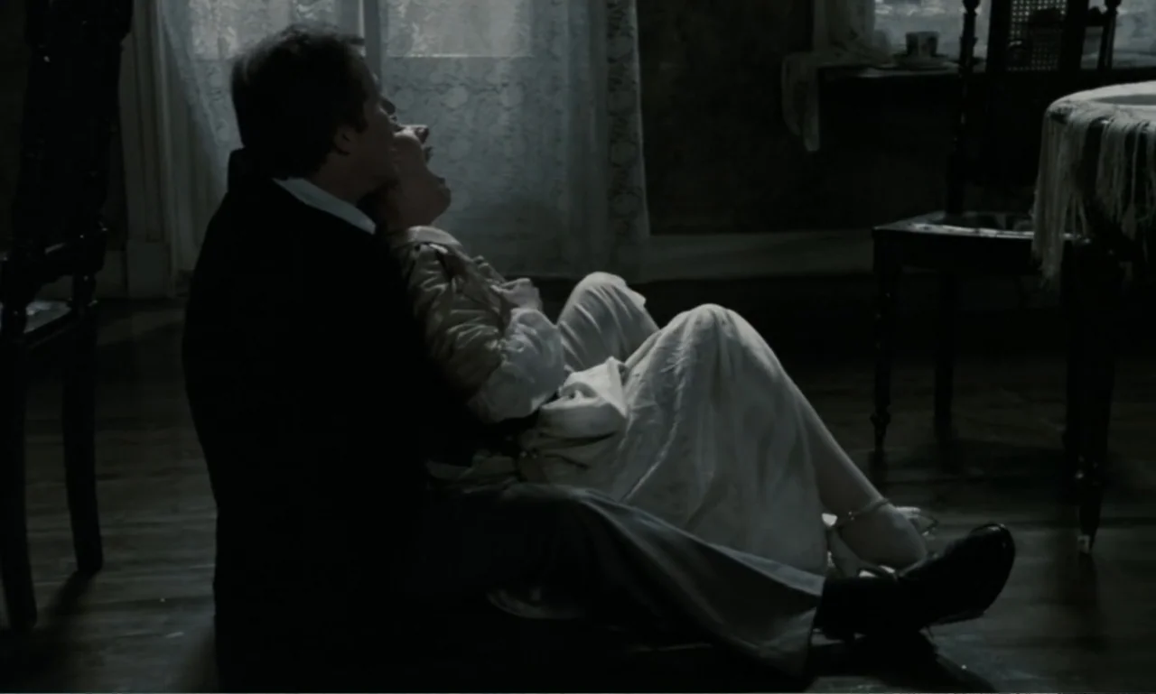 The Sacrifice (2015) movie still: medium — A dimly lit interior room features period furniture and a bright window with curtains. A man embraces a…