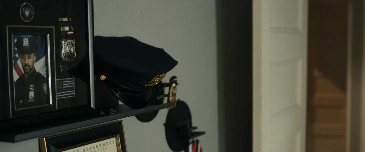 21 Bridges (2019) movie still: medium — A wall surface in a room features a black shelf holding law enforcement memorabilia. A framed photograph of…