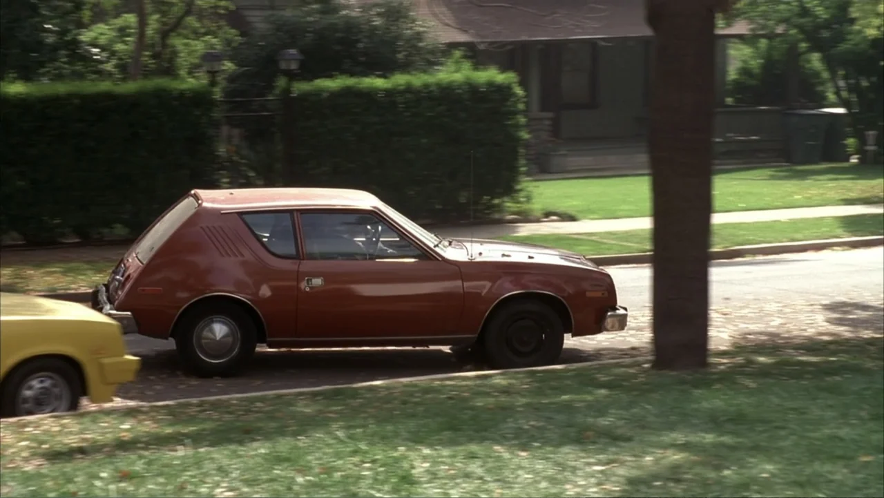 The Wedding Singer (1998) movie still: medium — A brown 1970s hatchback moves past a suburban house with a large manicured lawn. The car occupies…
