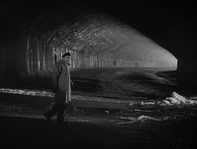 The Third Man (1949) movie still: wide — A dark subterranean concrete drainage tunnel contains a narrow pathway beside flowing water. A lone man in…