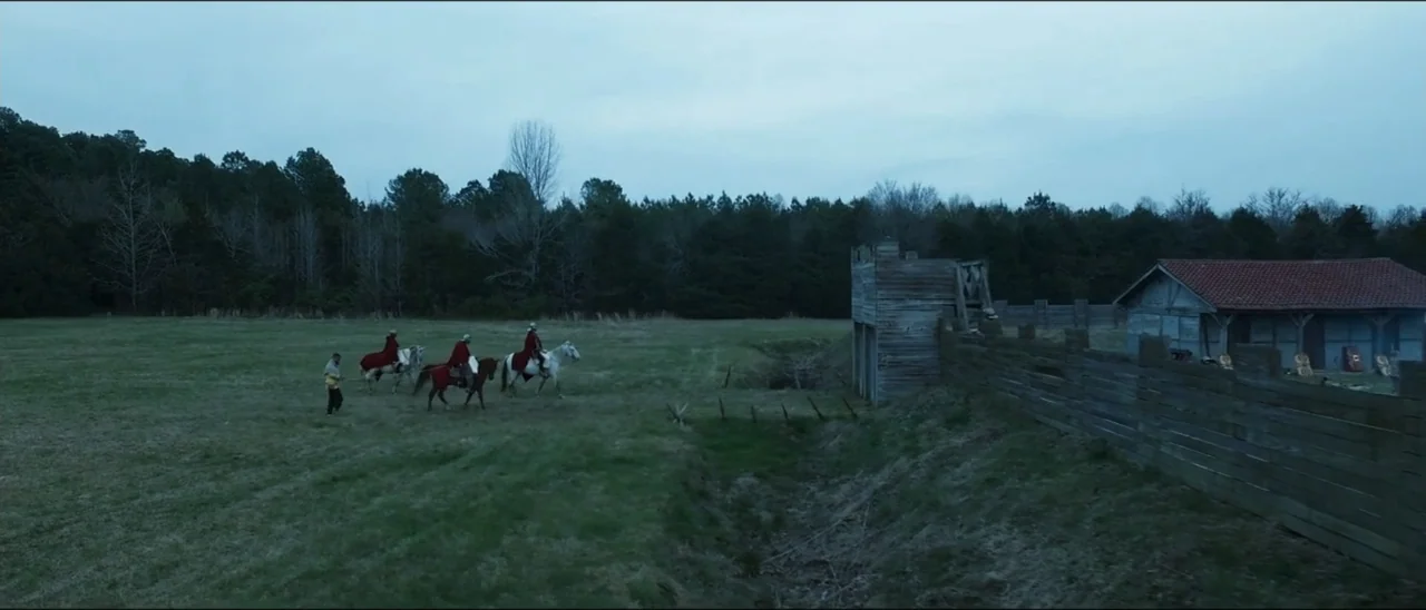 The Penitent Thief (2021) movie still: wide — An open grassy field contains a wooden watchtower and a simple roofed structure. Three riders wearing…