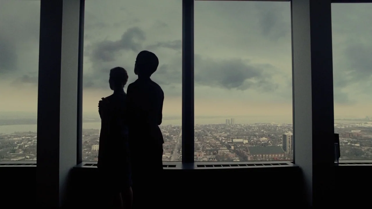 Splice (2009) movie still: fullbody — A high-rise room with floor-to-ceiling windows overlooks a sprawling urban landscape under clouds. A couple…