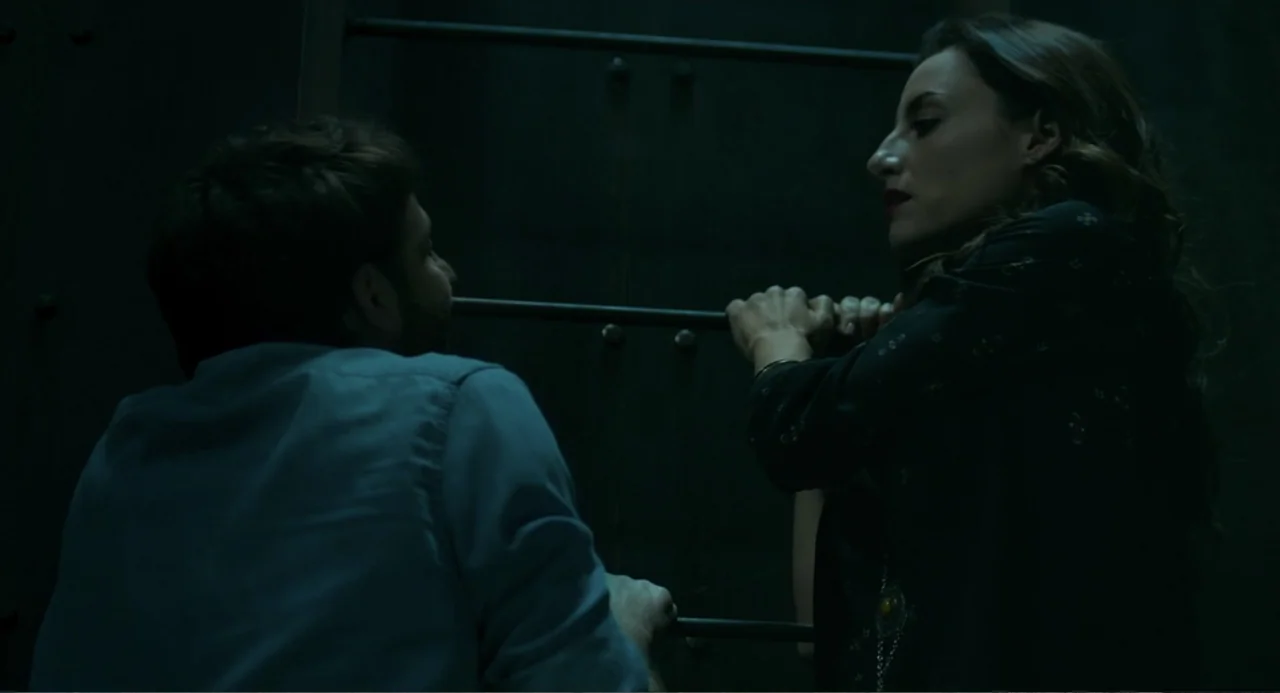 The Lift (2021) movie still: medium — Two individuals occupy a dark, confined metal elevator cabin with visible vertical bars. A man and a woman…