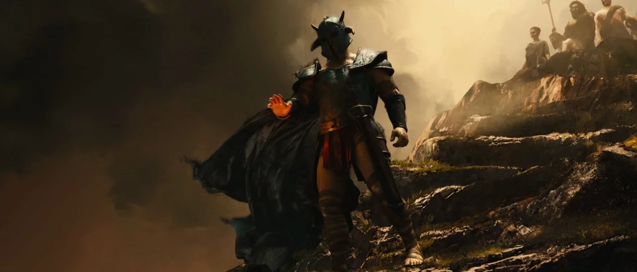 Wonder Woman (2017) movie still: fullbody — A warrior in dark metal armor stands atop a craggy, windswept rock formation. Observers watch from a…