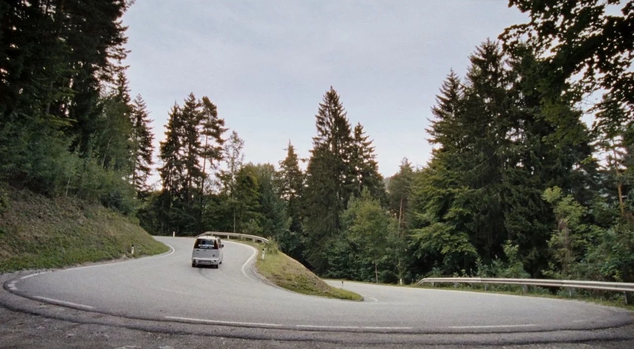 Paradise: Hope (2013) movie still: wide — A winding asphalt road curves through a dense coniferous forest with tall pine trees. A small,…