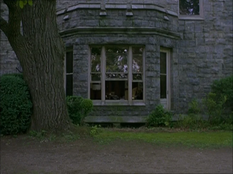 Last Days (2005) movie still: establishing — An exterior view shows the stone walls and multi-pane bay window of a house. A large dark tree trunk…