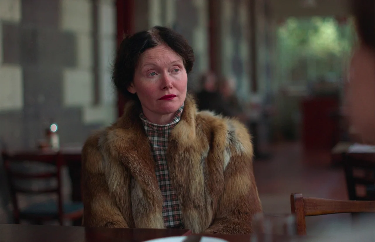 Nitram (2021) movie still: medium — A woman sits at a table inside a dimly lit eatery with dark wood furnishings. She wears a prominent fur coat and…