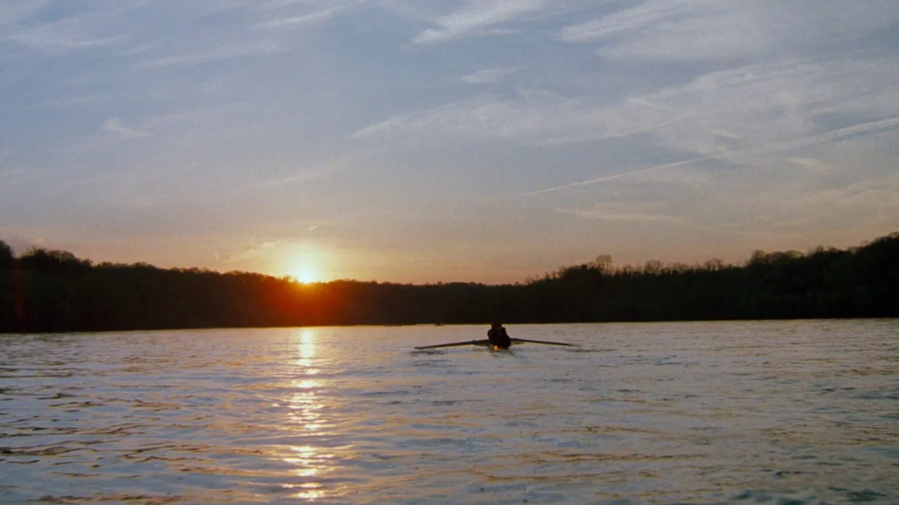 The Exorcist 3 (1990) movie still: wide — A calm lake reflects the light of a setting sun with tree-lined banks nearby. Two individuals are rowing a…