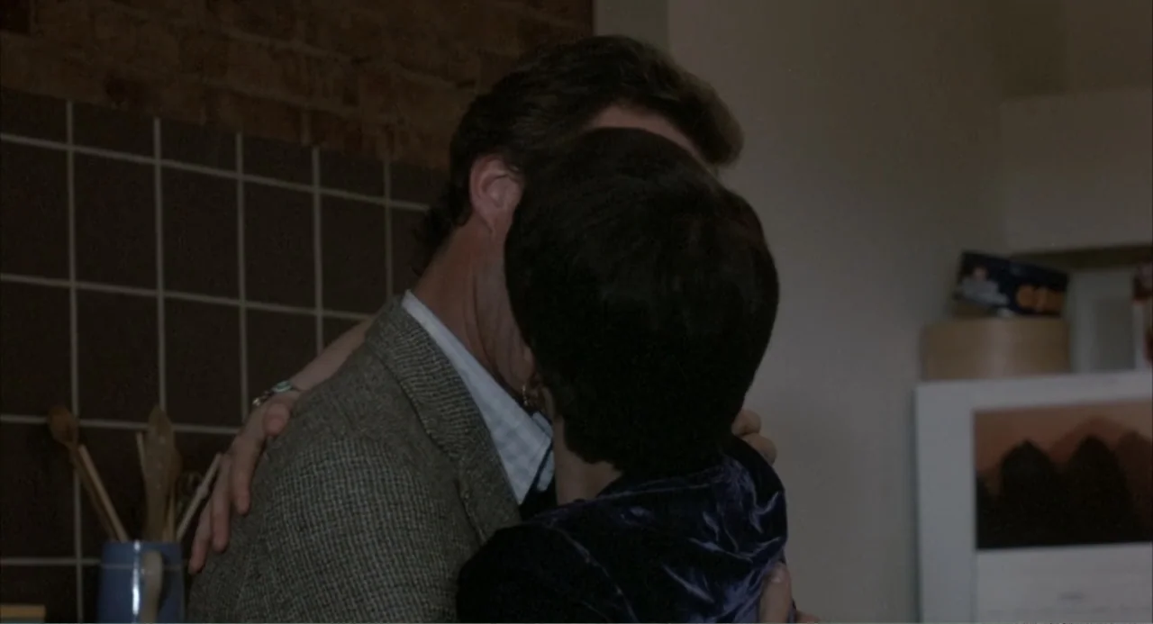 F/X (1986) movie still: medium — A residential kitchen features dark brown tiled walls and an exposed brick accent. A man in a tweed blazer and a…