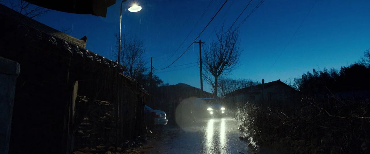 The Wailing (2016) movie still: establishing — A narrow rural road is bathed in the cool blue light of a night rain. A car approaches with…