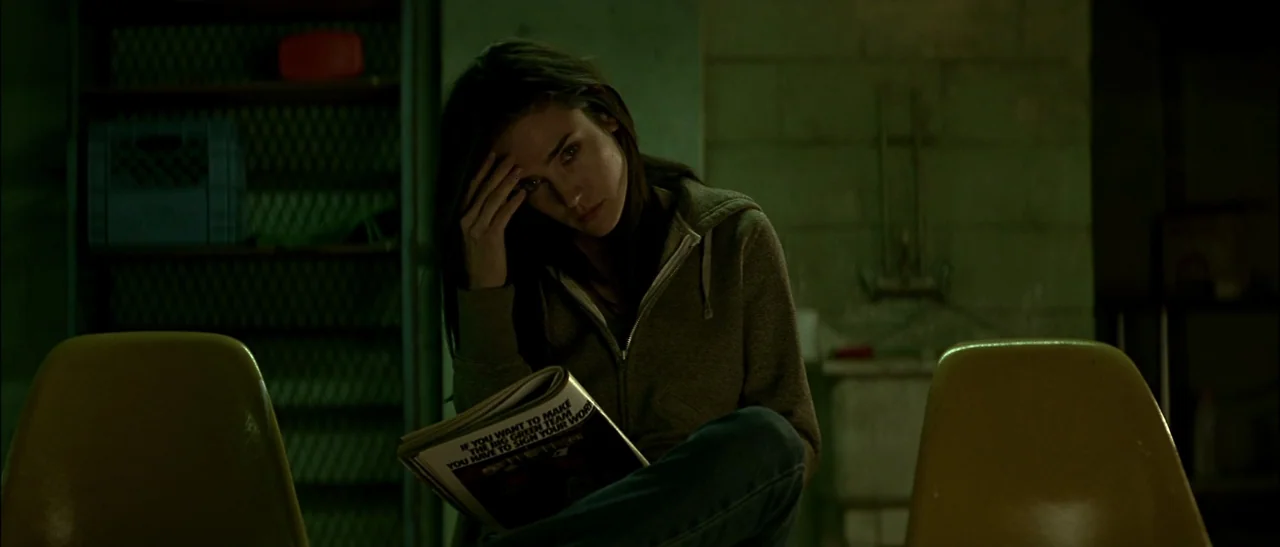 Dark Water (2005) movie still: medium — A dimly lit industrial-style waiting area features pale yellow plastic chairs and block walls. A woman with…