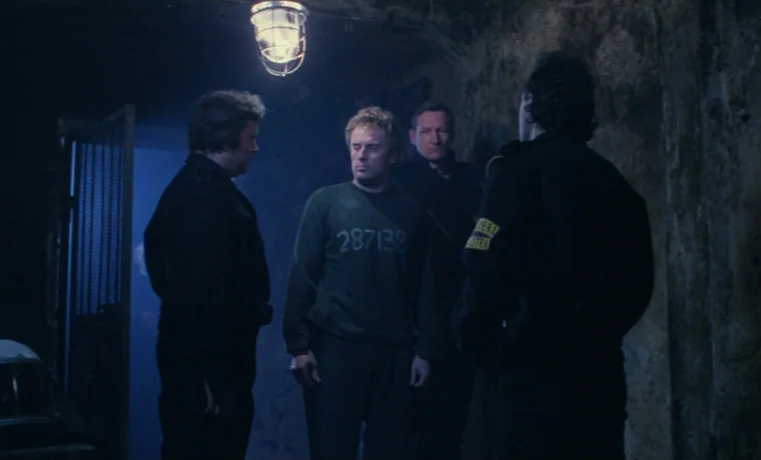 Ga-ga: Glory to the Heroes (1986) movie still: medium — A dimly lit narrow stone corridor with high walls and a metal grate door. Three uniformed…