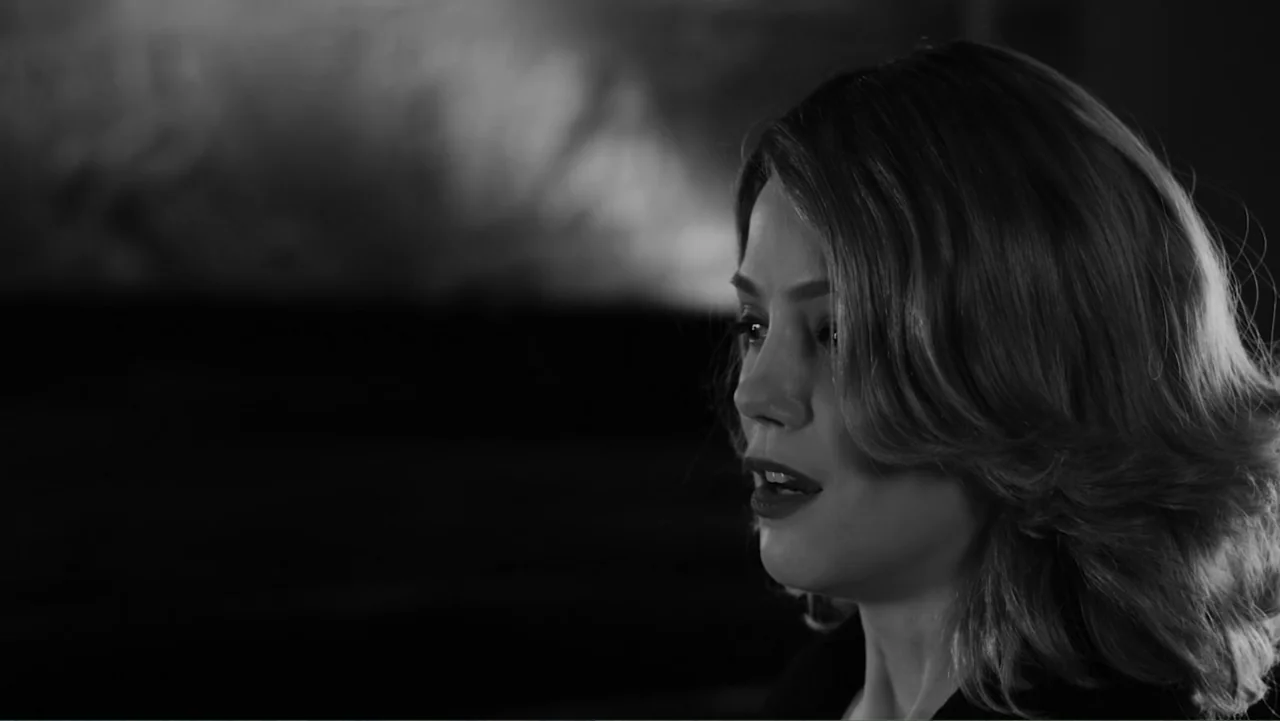 Creditors (2025) movie still: closeup — A dark interior space provides a stark backdrop for a close portrait of a woman. A woman with shoulder…
