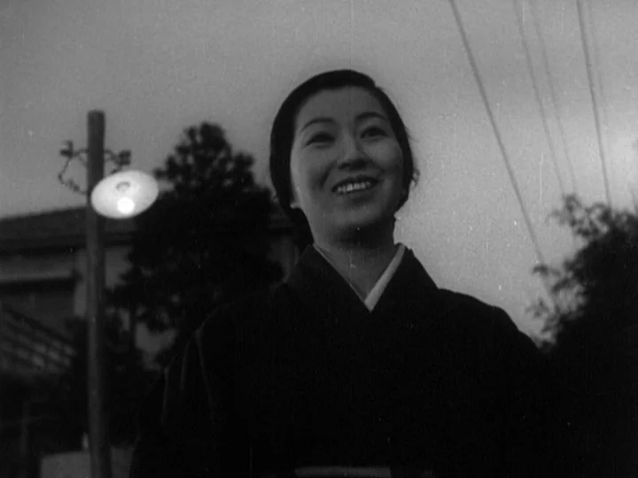 The Most Beautiful (1944) movie still: medium — An outdoor night setting featuring a woman standing in front of a pole lamp. She looks away from the…