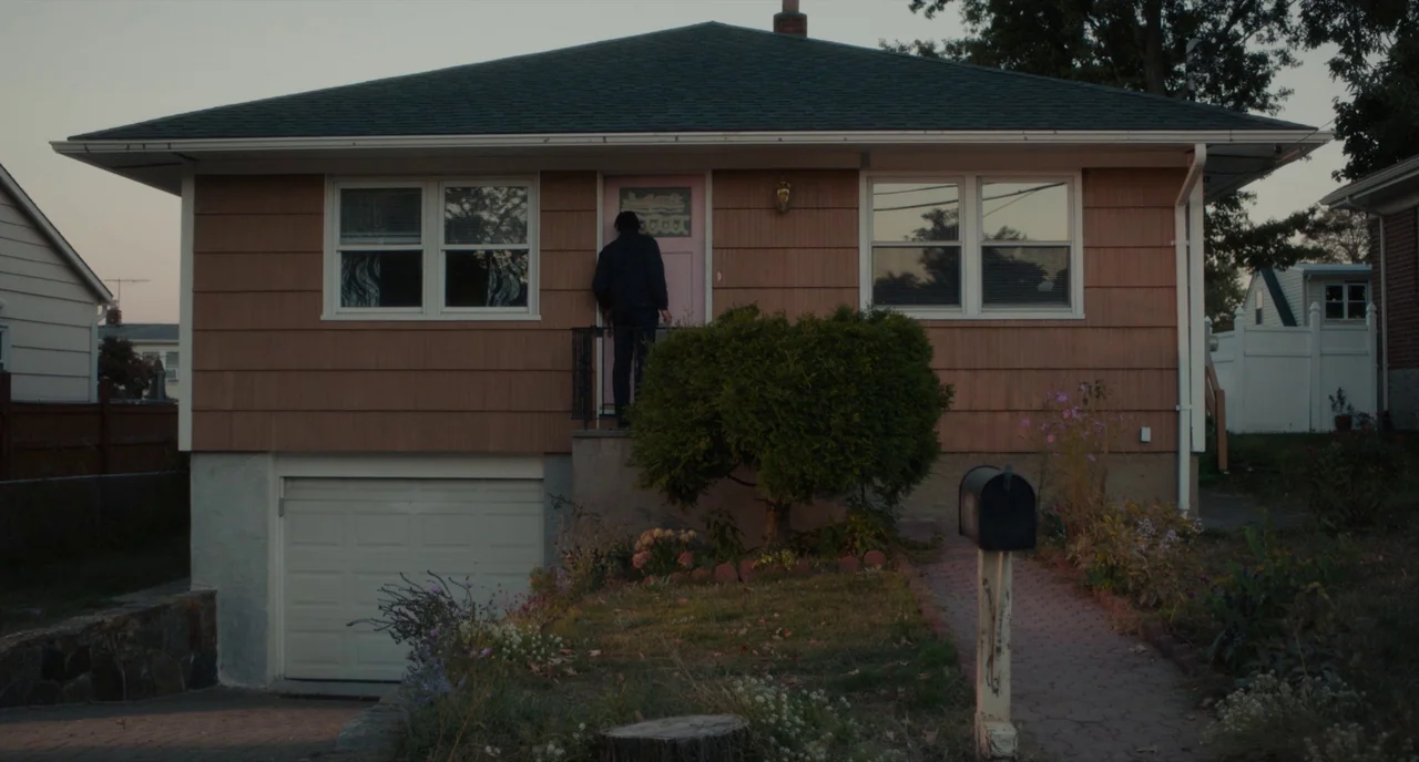 Paterson (2016) movie still: wide — A split-level residential house has tan horizontal siding and a dark shingled roof. A person stands on the small…