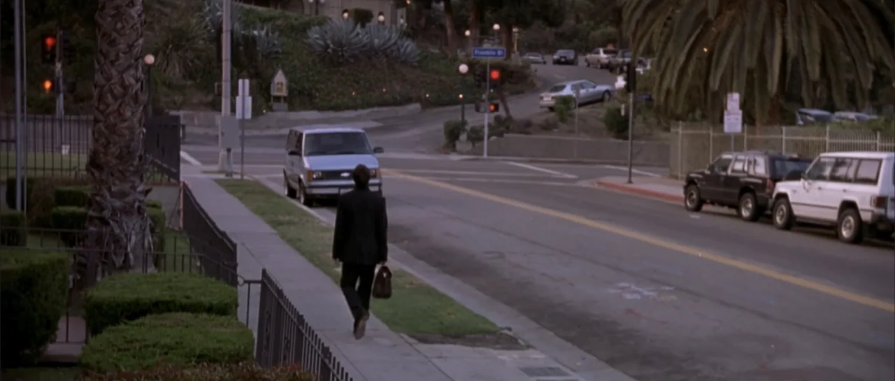 Shade (2003) movie still: wide — An urban intersection with parked vehicles and landscaping under a dusky sky. A lone man in a suit walks away along…