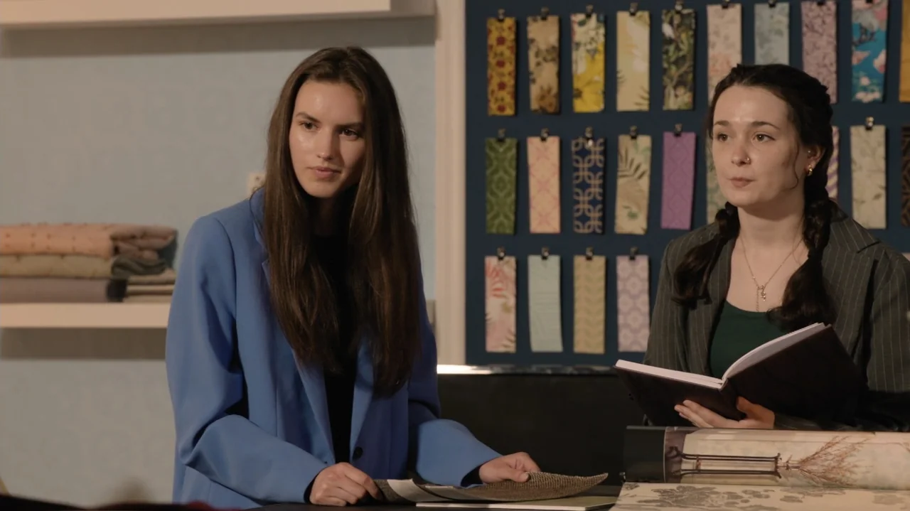 Designing Christmas (2022) movie still: medium — A design studio wall features vertical fabric samples mounted on a blue display board. Two women in…