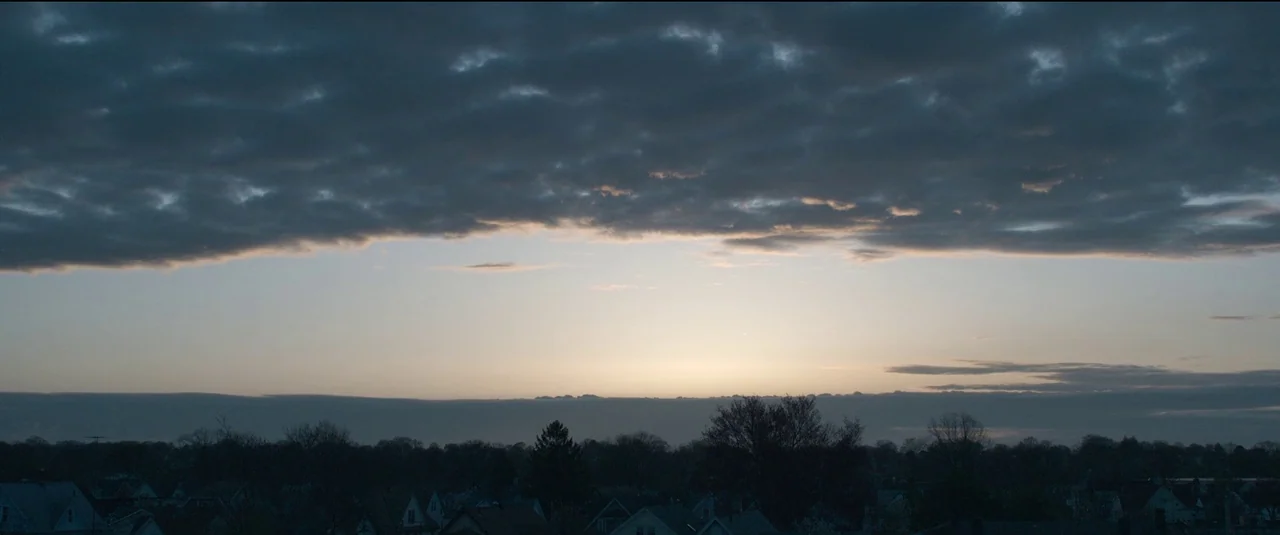 The Good Nurse (2022) movie still: establishing — An expanse of gray storm clouds hangs over a dark, silhouetted suburban skyline. The pale light of…