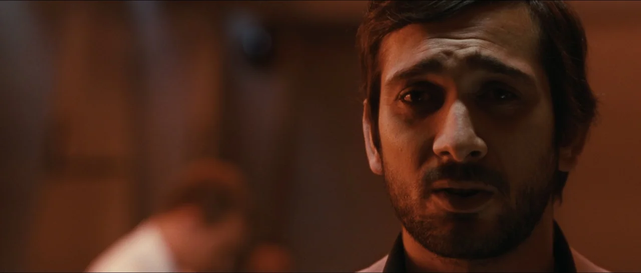 Slackers (2002) movie still: closeup — A dimly lit room with an out of focus background. A man with dark stubble looks toward the camera with his…