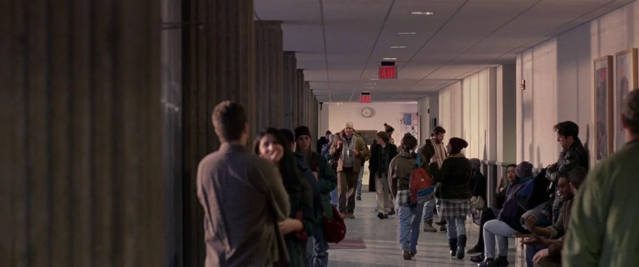 The End of the Tour (2015) movie still: wide — A long, institutional school hallway features columns, tiled floors, and rows of lockers. Students…